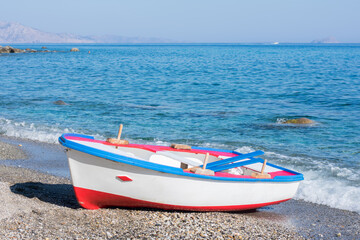 Colorful painted wooden boat on the shore of a stony beach with calm blue water in Ikaria, Greece.