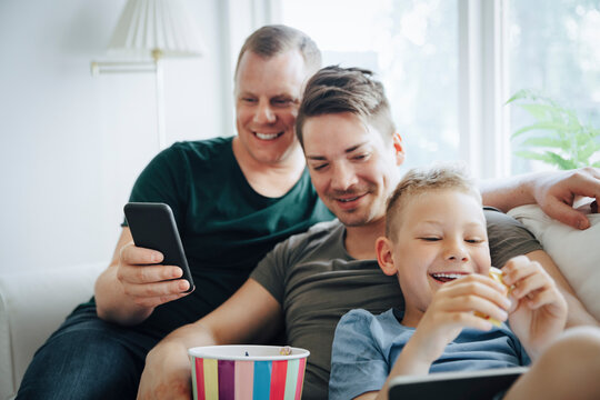 Smiling Fathers And Son Using Technology While Eating Popcorn On Sofa At Home