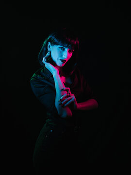 Girl with black hair and black makeup posing on a black background with colored light. She demonstrates her hands and stares at the camera.