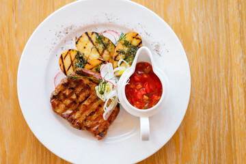 Fried  barbeque meat on a white plate with potatoes and vegetables. Grilled BBQ decorated with vegetables and sauce on wooden table and parchment paper.