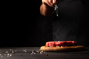 cook sprinkles salt on a piece of meat on a black background. The hand of the cook is in the frame. Horizontal photo with free space for text logos and advertisements on the left side.