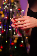 A girl with a gold ring on the middle finger holds a glass of champagne close-up against the background of a New Year tree with colorful lights. Christmas decor.