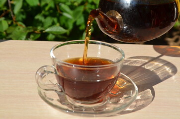 A girl pours tea from a transparent teapot into a transparent cup against a background of green foliage Good morning, outdoor cafe, energy boost. Tea being poured into glass tea