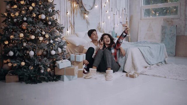 Young couple hugging and taking pictures with the phone, while sitting next to the Christmas tree at home. Bright living room decorated with bright Christmas tree lights. Happy winter holidays and
