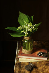 On a wooden background, a vase with a bouquet of white lilies of the valley standing on books and chocolate milk.

