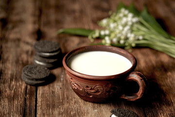 There is a bouquet of lilies of the valley on a wooden table, milk and chocolate chip cookies in a clay cup.
