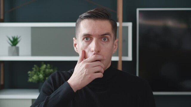 A surprised young millennial men with brown hair green eyes in black sweater is opening her mouth at a loss and looking to the camera on modern interior background. 4k