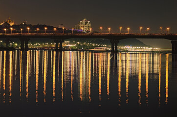 Beautiful landscape photo of Kyiv at night. Famous Paton Bridge over Dnipro River. Mirror reflections on smooth water of the river. Long time exposure. Beautiful autumn evening. Kyiv, Ukraine