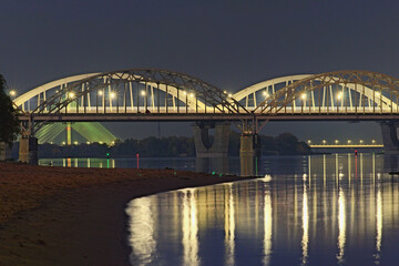 Three illuminated bridges at night. Picturesque landscape of Dnipro River with arched Railway bridge, Darnytskyi Rail and road Bridge. Pivdennyi Bridge in the background. Scenic evening landscape