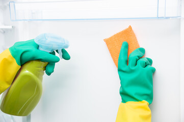 Close-up of woman's hand wearing yellow gloves cleaning open refrigerator with spray bottle and...