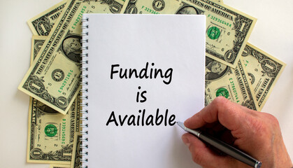 Male hand writing 'funding is available' on white note, on white background. Dollar bills. Business concept, copy space.
