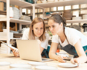 Two female entrepreneur with laptop in artisan workroom. Waist up portrait of cheerful business woman artisan in pottery studio. Concept for lady in freelance