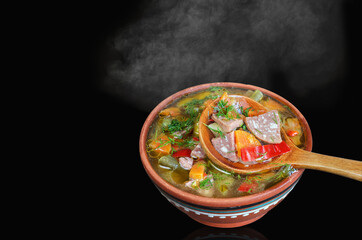 A bowl of hot soup with vegetables and sausage, steam rises. Close-up in a spoon