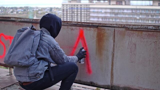 Process of making anarchy graffiti by young protester in a hood with red spray paint