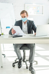 Full length of businessman in medical mask looking at paper, while sitting at table in office on blurred background