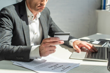 Cropped view of sad businessman with credit card, typing on laptop, while sitting at workplace on blurred background