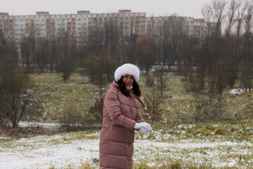 Fototapeta premium A girl in winter clothes on a walk in the park. Sculpts a ball of snow with his hands. The ground is covered with the first snow