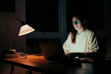 Young female entrepreneur sending emails sitting at office desk working late at night in apartment...