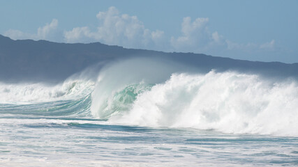 Breaking waves against the backdrop of a ridge and a cloudy sky. The beach is located on the North Shore of the Island of Oahu.