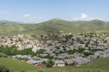 Wide view of a hillside town with houses and green rolling hills under a blue sky in western Iran.
