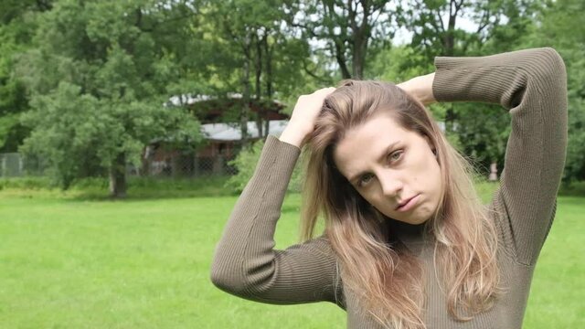 Beautiful young blonde girl walks in a summer green park, posing, looking at the camera and straightens her hair