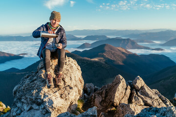 Active backpacker hiker woman sitting on the mountain summit cliff, pouring a tea from a thermos flask and enjoying mountains valley covered with clouds view. Successful summit climbing concept image.