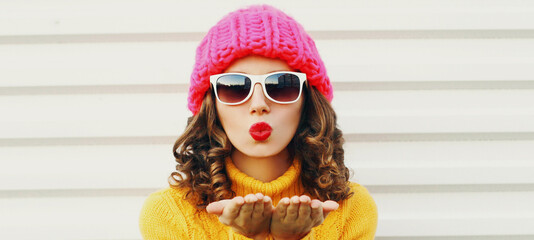 Winter portrait close up of young woman blowing red lips sending sweet air kiss wearing a pink hat,...