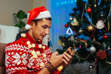 Man using smartphone for Christmas. Guy celebrating New year alone at home wearing Santa's hat, lights by tree.