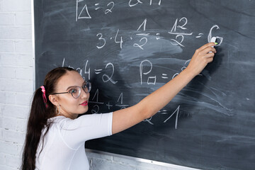 A teenage girl is solving a math problem at school by writing on a school blackboard with white chalk. Her hair is pinned with a pink ribbon in two ponytails.