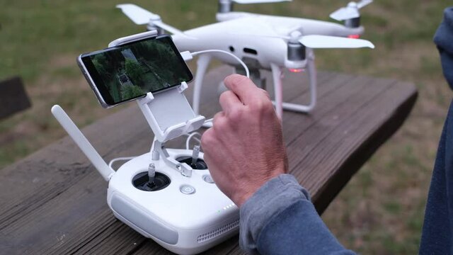 Drone pilot checking videos on smartpohone after a flight. Hand with white rc controller, close-up video with a quadcopter in the background. Flying drones, aerial photography or videography concept.