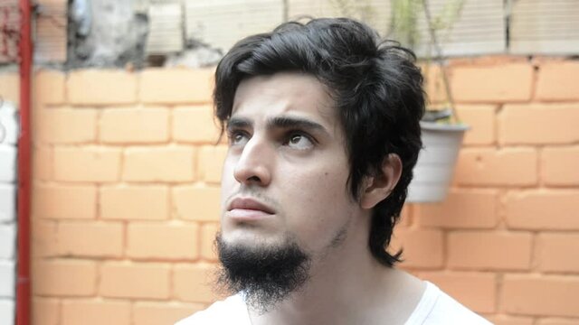 Latino male model in white shirt looking to the side and up, donning a light blue medical mask with a orange brick wall in the background. Scene of new normality during the covid 19 pandemic