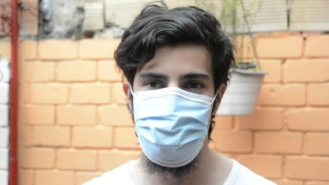 Latino male model in white shirt looking to the camara smiling and donning a light blue medical mask, with a orange brick wall in the background. Scene of new normality during the covid 19 pandemic