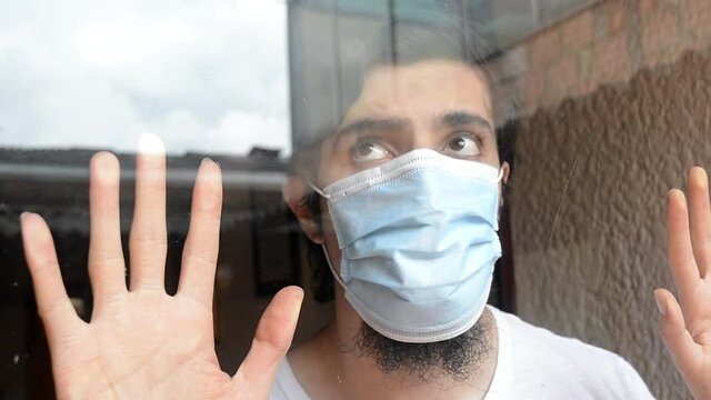 Latino male model in white shirt and light blue medical mask looking through the window with a cloudy sky and buildings reflexed. Scene of new normality during the covid 19 pandemic