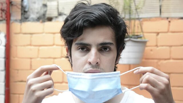 Latino male model in white shirt looking to the camara and donning a light blue medical mask, with a orange brick wall in the background. Scene of new normality during the covid 19 pandemic