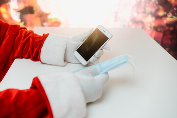 Santa Claus hands holding smart phone and a medical mask over white table background. Online greetings, ordering services for Christmas and new year.