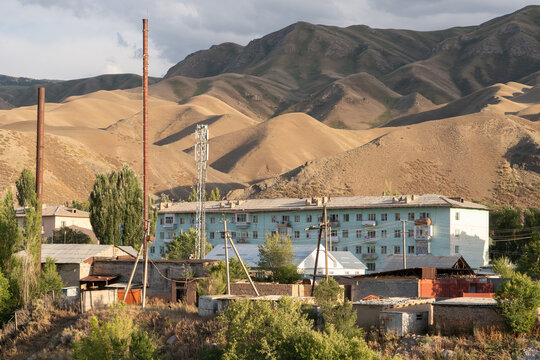 Village scene in Naryn, Kyrgyzstan, with a multi-storey building, houses and wooden power poles set against brown foothills under a partly cloudy sky.