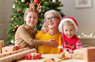 happy family grandma and grandkids hug and smile for camera while wrapping Christmas gifts near a...