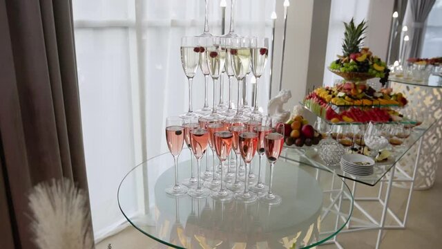 A three-story slide made of thin glass glasses filled with white and pink champagne and with bunches of berries stands on a stand and complements the sophistication of an expensive and elegant buffet