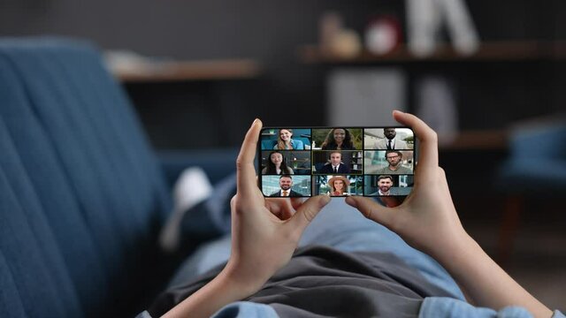 The woman is lying, relaxing on a Coach in the Living Room, holding a Smartphone in Horizontal Mode. The woman is Watching Seminars, Webinars with a Group of Diverse People. Online Surfing Lifestyle.
