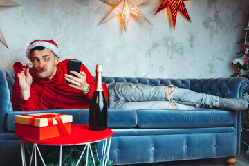 Young handsome man in Santa Claus hat with glass of champagne taking selfie on smartphone. Funny guy photographing himself on mobile phone and lying on couch. Concept of Christmas celebration at home.