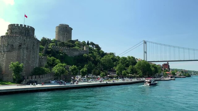 Footage of a yacht passing on Bosphorus strait and people walking at upscale neighborhood called "Rumeli Hisari". Historical fortress and FSM bridge are in the view. It is a beautiful Istanbul scene.