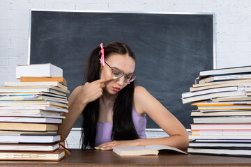 A teenager sits in the classroom among the piles of books and begins reading the first reading. The student starts learning. Her hair is pinned with a pink ribbon in two ponytails.