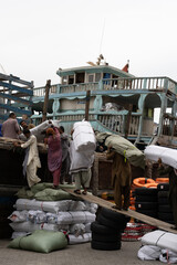 Traditional wooden cargo dhow moored at the quay along Dubai Creek in the United Arab Emirates, as men load sacks and goods onto its deck. Weathered multi-storey buildings rise behind the bustling sce