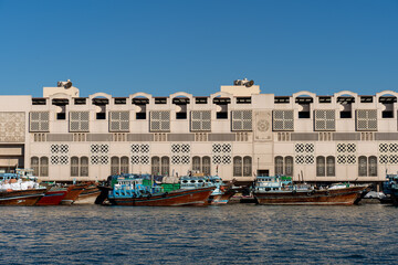 Traditional wooden dhows moored in front of the Deira waterfront in Dubai, United Arab Emirates. The beige concrete facade with repetitive arches reflects in the calm creek water under a bright Januar