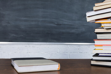 A pile of books stacked on either side of a school desk. One book is in the center of the table top. There is a clean blackboard in the background.