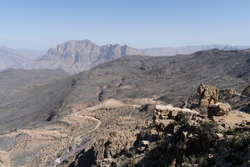Wide-angle view of a rugged mountain canyon with a winding road descending into the Al Hajar Mountains of northern Oman. Sheer cliffs and rocky slopes frame the deep valley under a hazy blue sky in Fe
