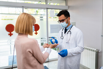 Young male practitioner with face protective mask working at clinic reception desk. He is holding disinfectant bottle and helping his patient to disinfect hands.