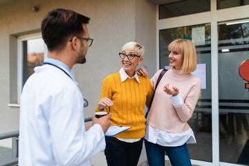 Obraz premium Young male practitioner or doctor talking with middle age female visitor and her mother in front of medical clinic or nursing center.