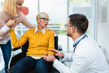 Obraz premium Young male practitioner working at medical clinic reception desk and helping to middle age woman and her sick mother which sitting in disability chair.
