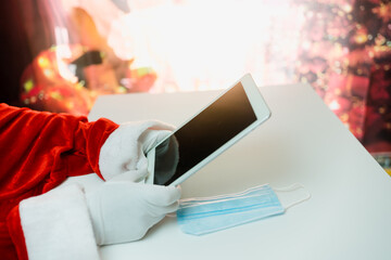 Fototapeta premium Santa Claus hands holding tablet and a medical mask over white table background. Online greetings, ordering services for Christmas and new year.